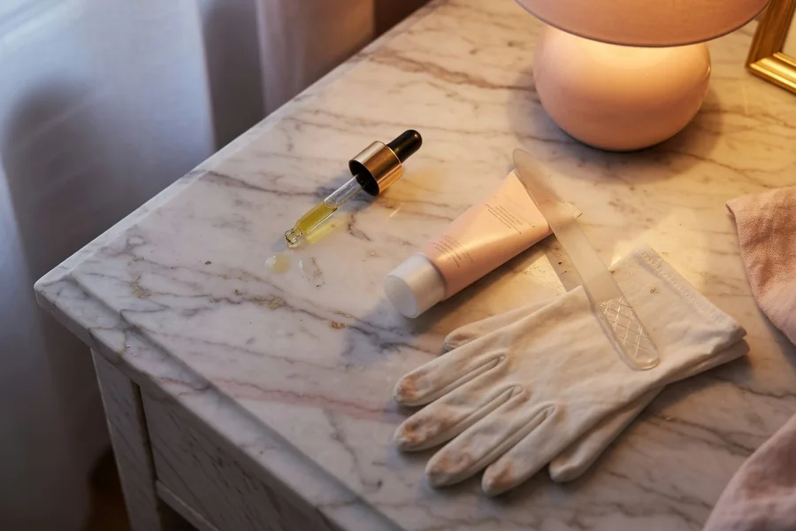 Cuticle oil, hand cream, cotton gloves and a glass file on a bedside table