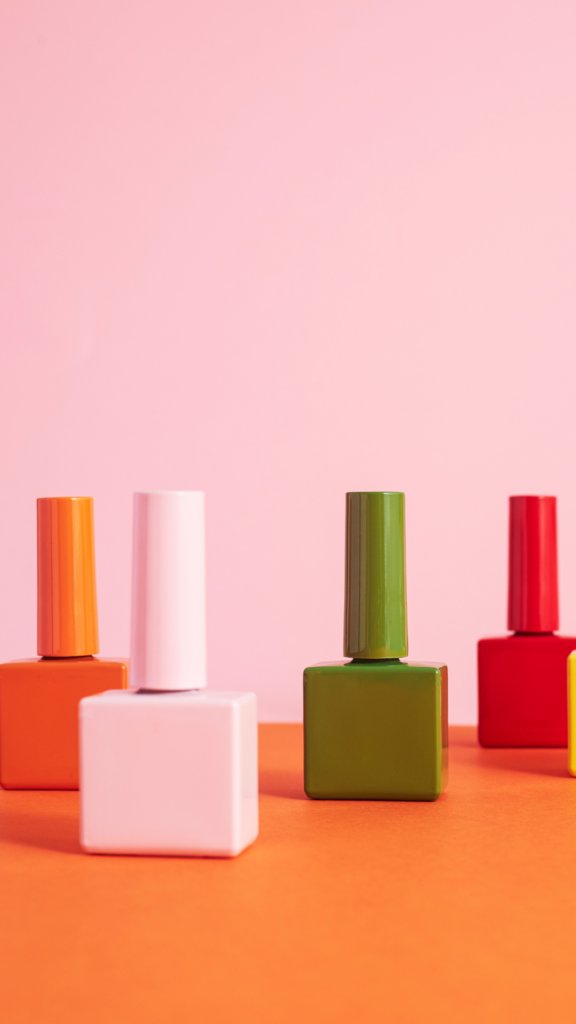 tendance vernis de 2025 Des flacons de vernis à ongles colorés, aux formes carrées et aux bouchons allongés, sont disposés sur une surface orange avec un fond rose pastel.
